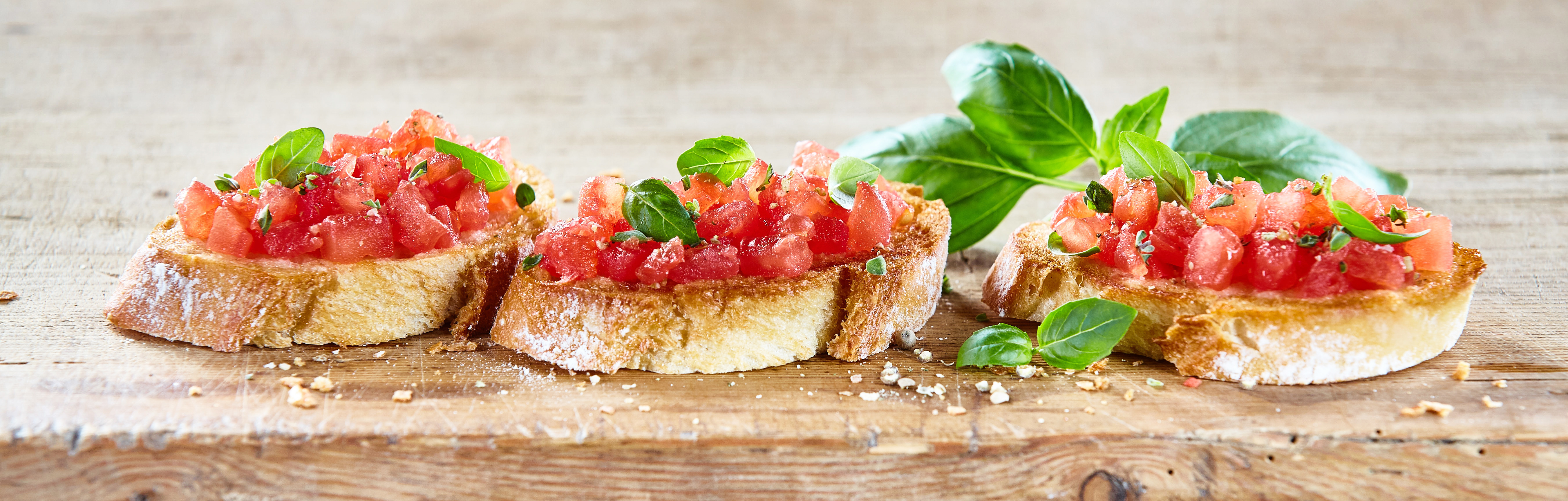 Delicious seasoned savory Italian tomato bruschetta slices on a rustic wooden board garnished with fresh basil leaves, horizontal banner